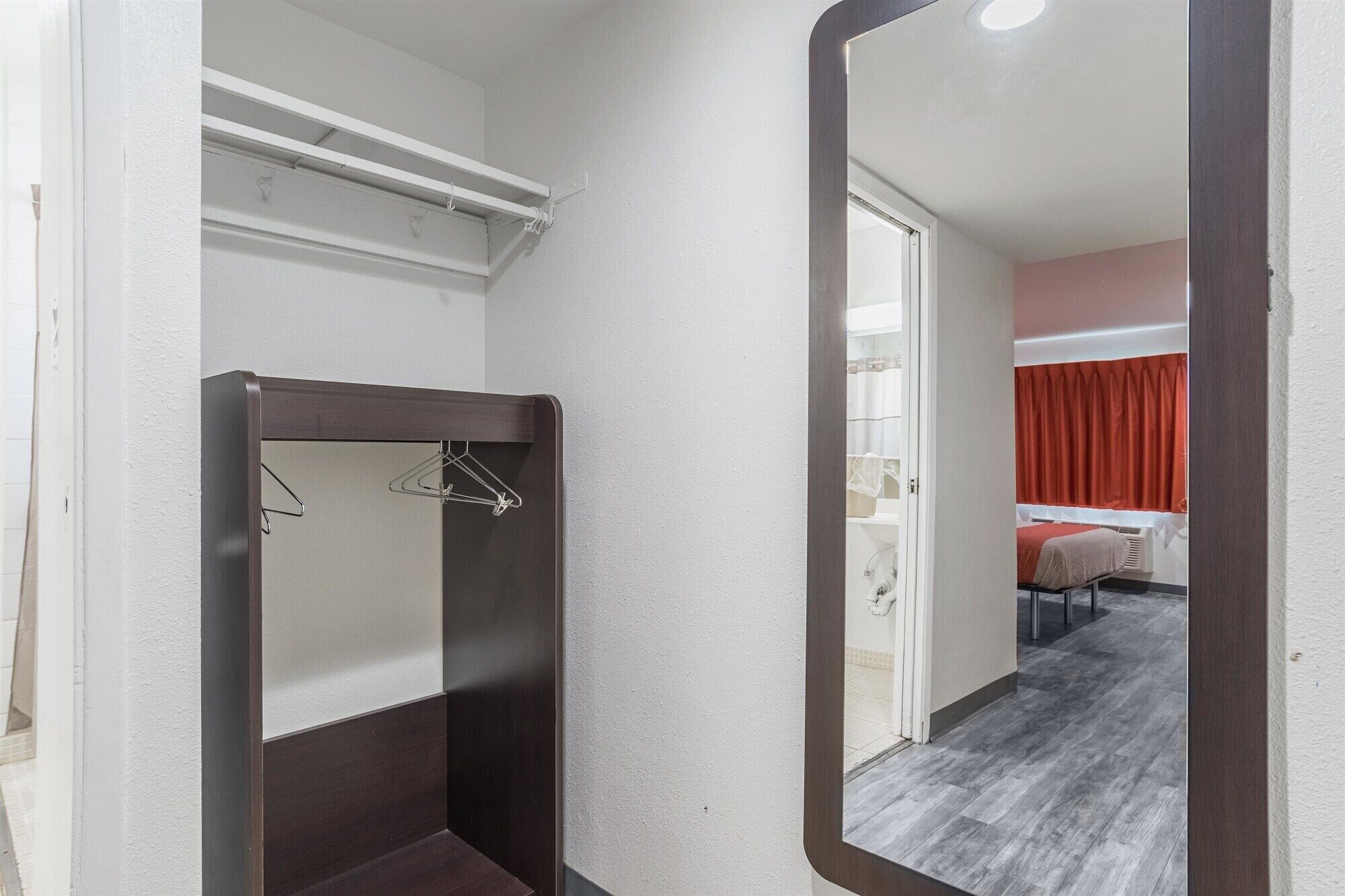 A hotel room with a dark wood vanity, a mirror, and a view of a bed with red curtains.