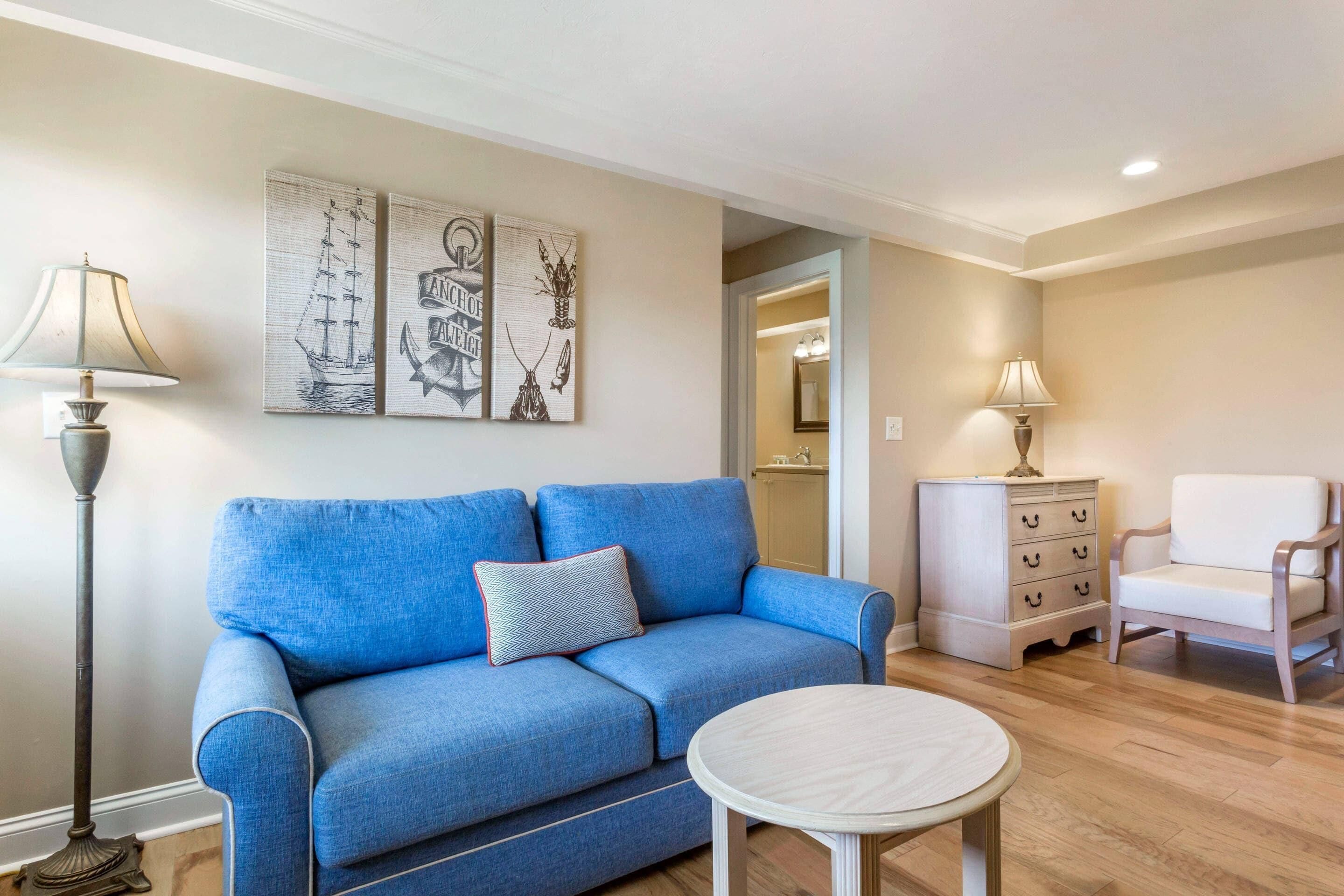 Una sala de estar con un sofá azul, una mesa redonda, un sillón blanco, una lámpara de pie y un aparador.