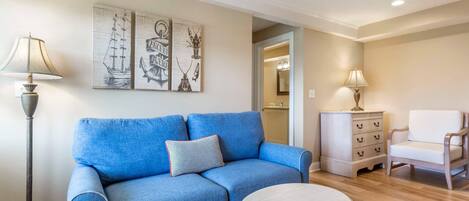 Una sala de estar con un sofá azul, una mesa redonda, un sillón blanco, una lámpara de pie y un aparador.