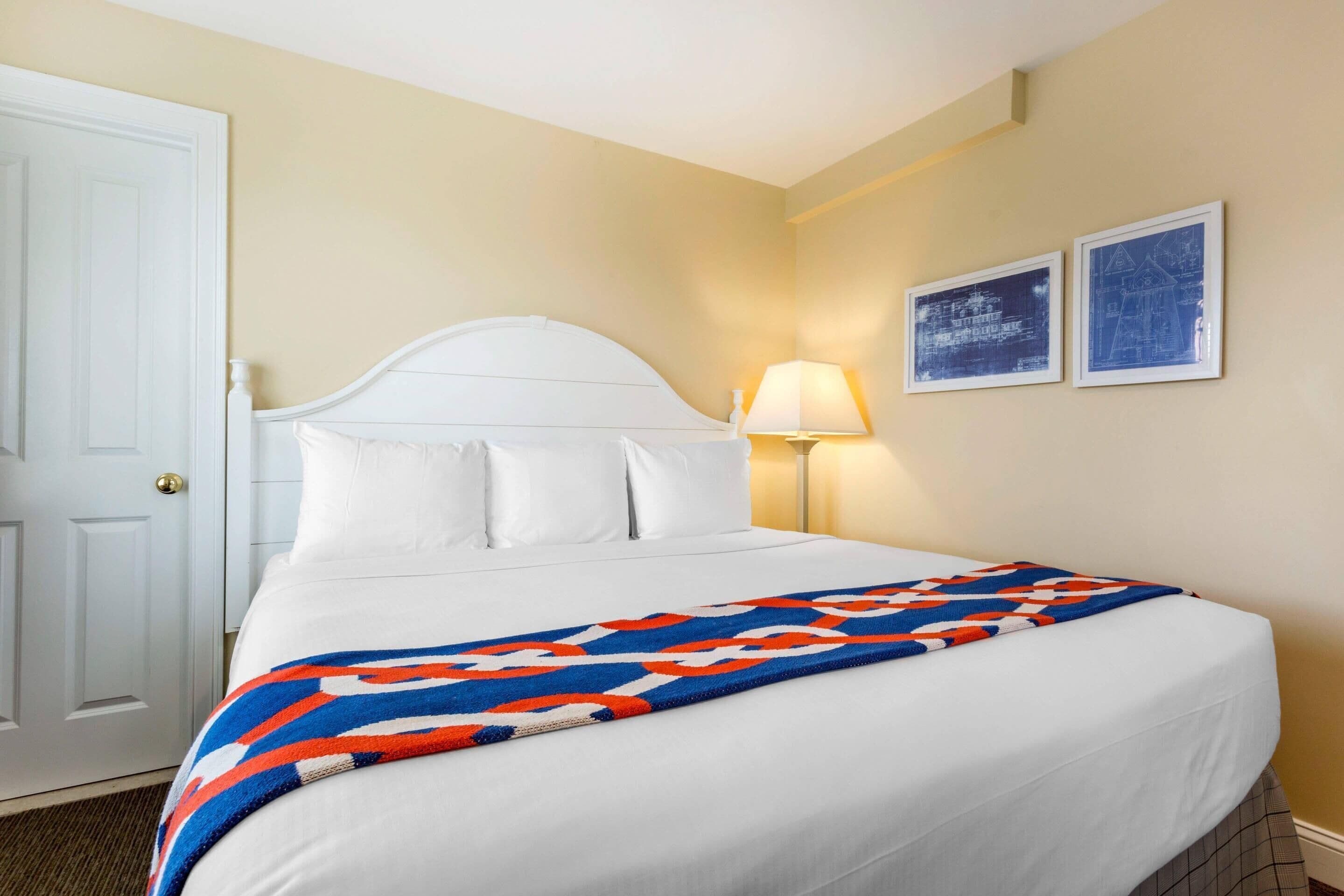 Een hotelkamer met een wit bed, een kleurrijk kleed, een witte deur en twee ingelijste foto's aan de muur.