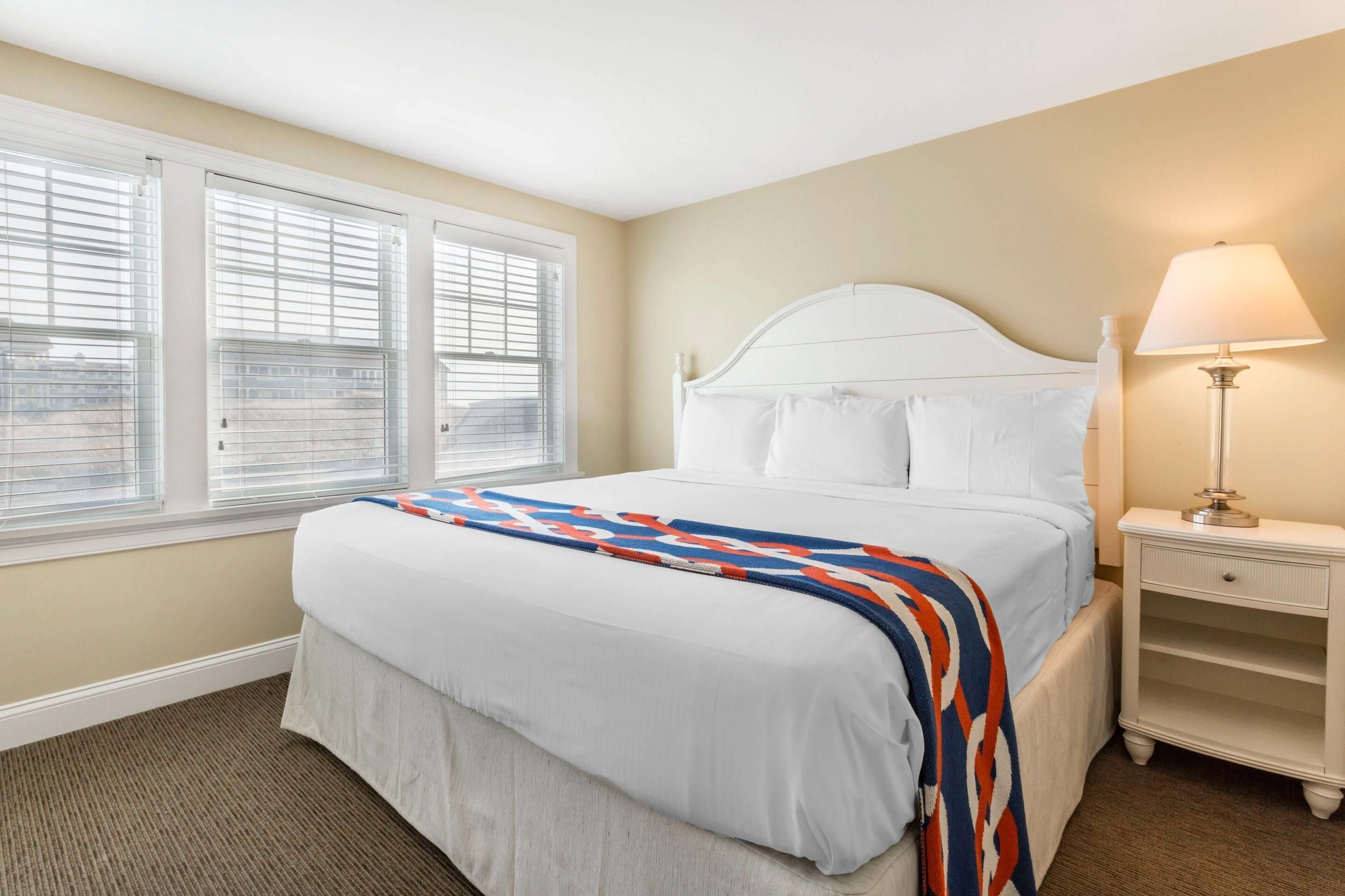 A neatly made bed with a white headboard, a bedside table with a lamp, and a window with blinds.