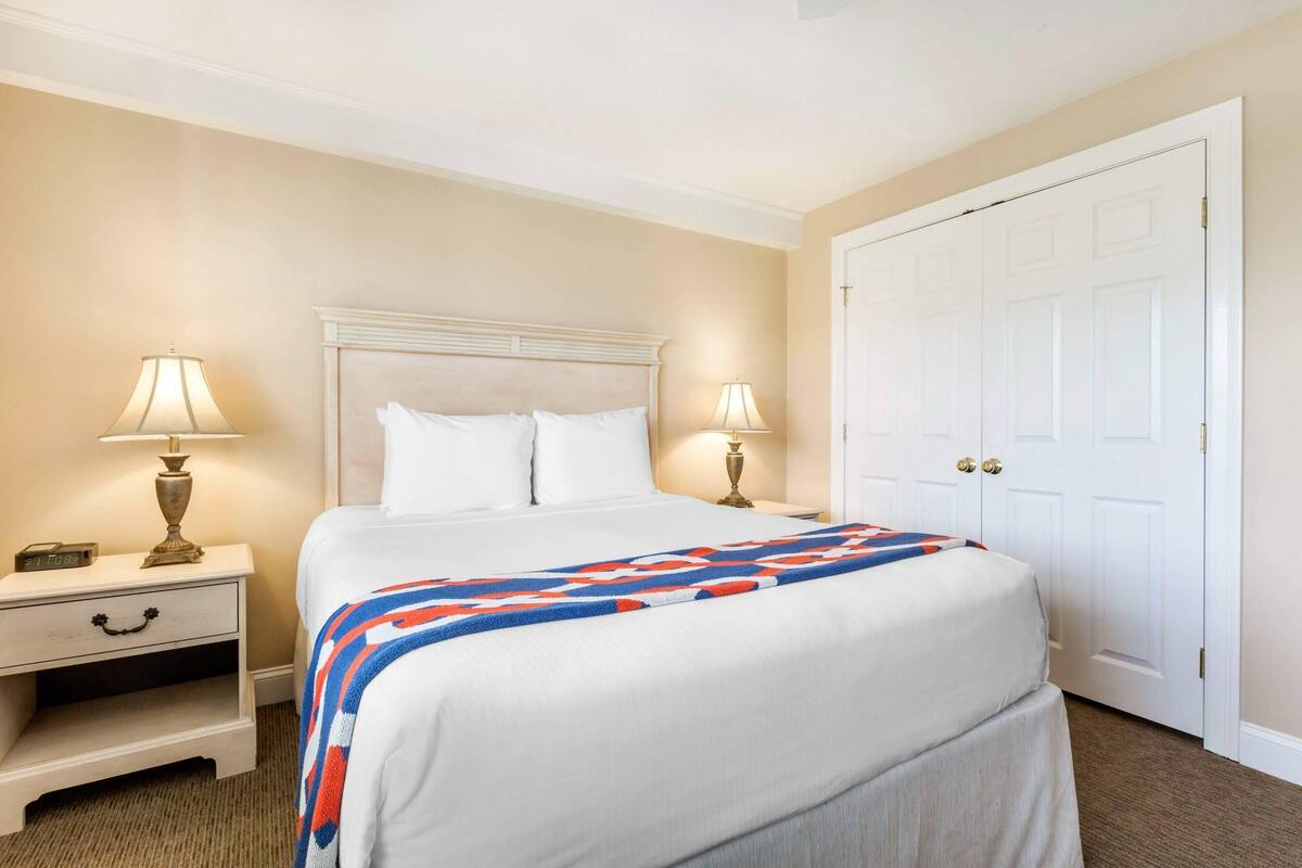 A hotel room with a large bed, two bedside tables with lamps, a white door, and a carpeted floor.
