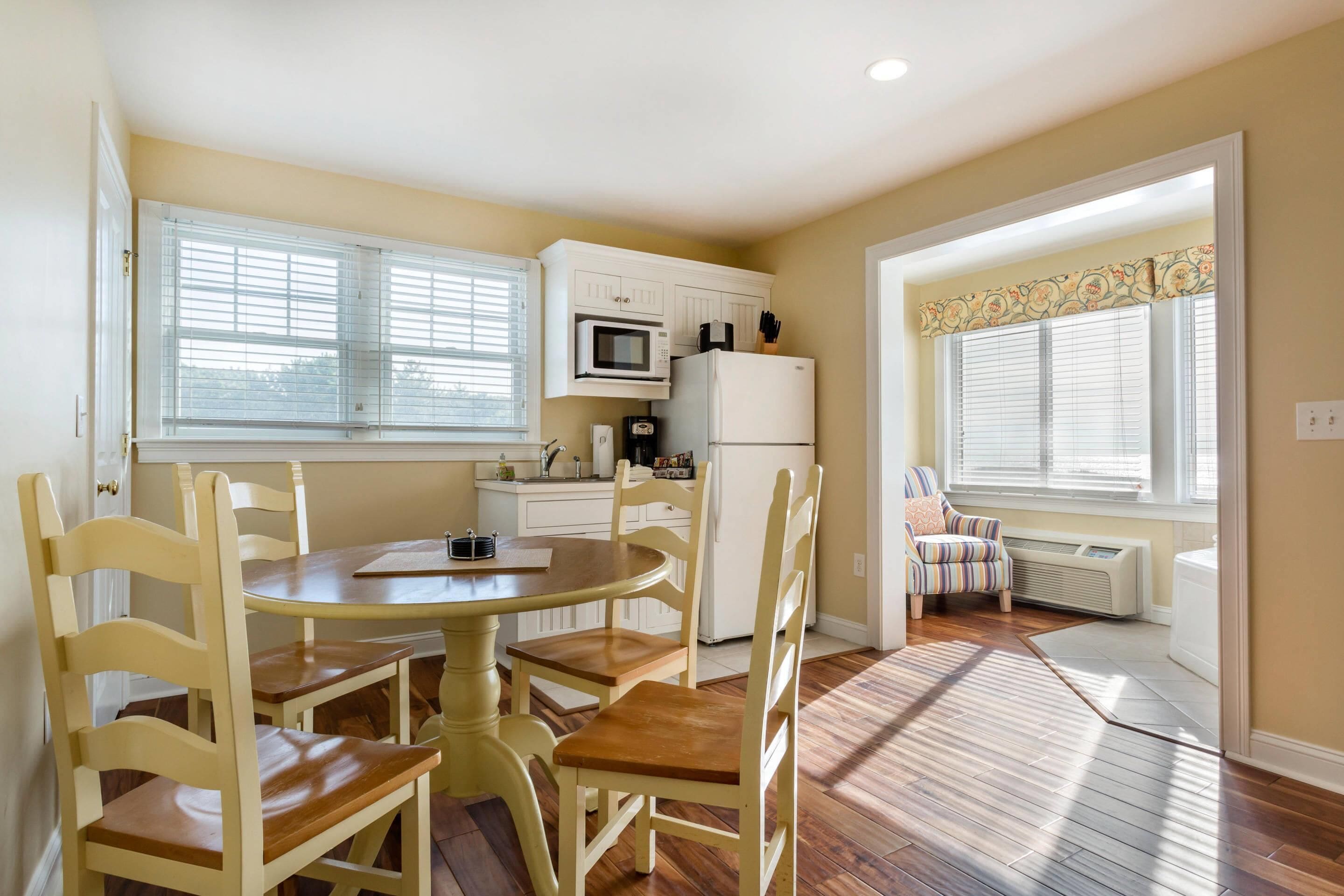 A kitchen with white cabinets, a microwave, and a refrigerator. There is a dining table with chairs and a window with blinds.