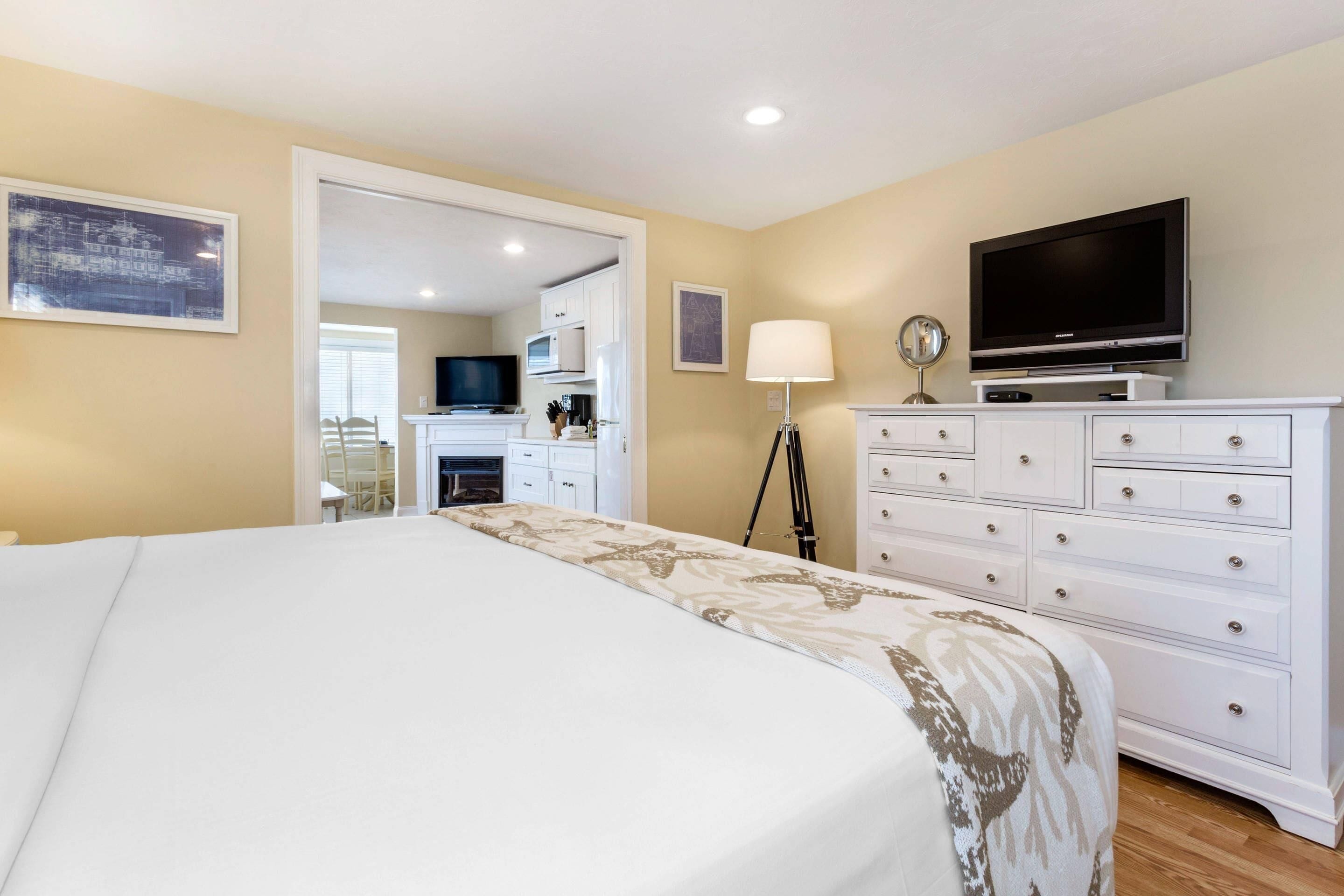 A bedroom with a large bed, a TV on a dresser, a lamp, and a view into the kitchen.