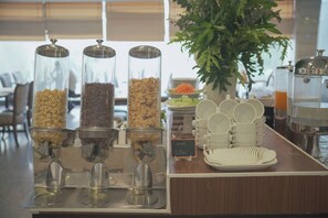 Mesa de buffet con opciones de cereales y granola, una máquina de café y una planta al fondo.