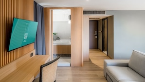 Une chambre d’hôtel moderne avec un mur lambrissé en bois, un téléviseur à écran plat, un canapé, un coin repas et une salle de bain équipée d’un lavabo et d’une baignoire.