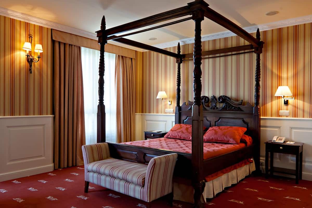 A four-poster bed with a red bedspread, a striped armchair, and two bedside lamps.