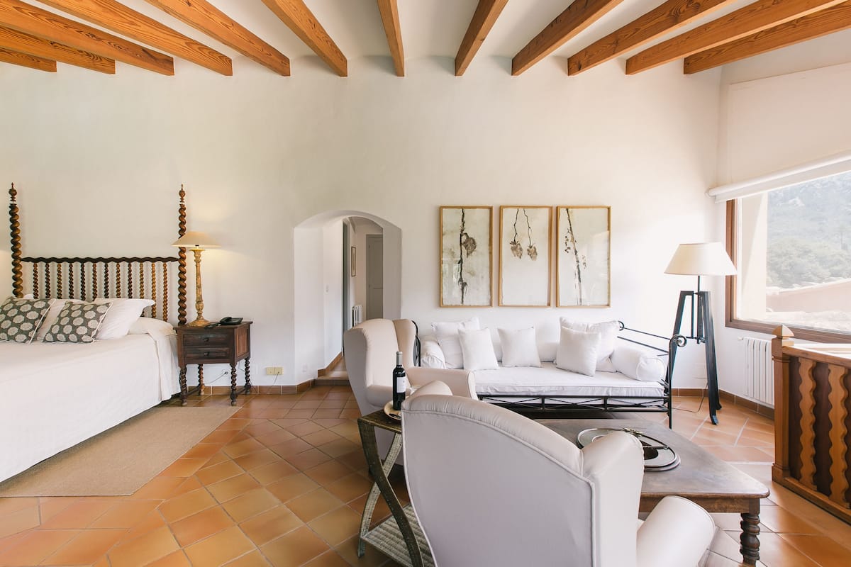 A spacious living room with a sofa set, a coffee table, and a dining table. There is a wooden ceiling with exposed beams, a white wall with framed artwork, and a window providing natural light.