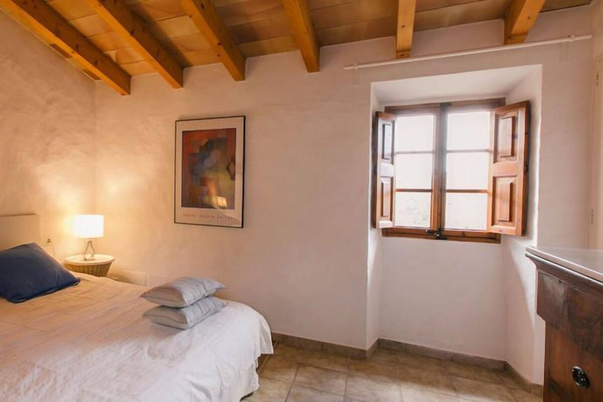 A bedroom with a bed, pillows, a nightstand with a lamp, a framed picture, and a window with wooden shutters.