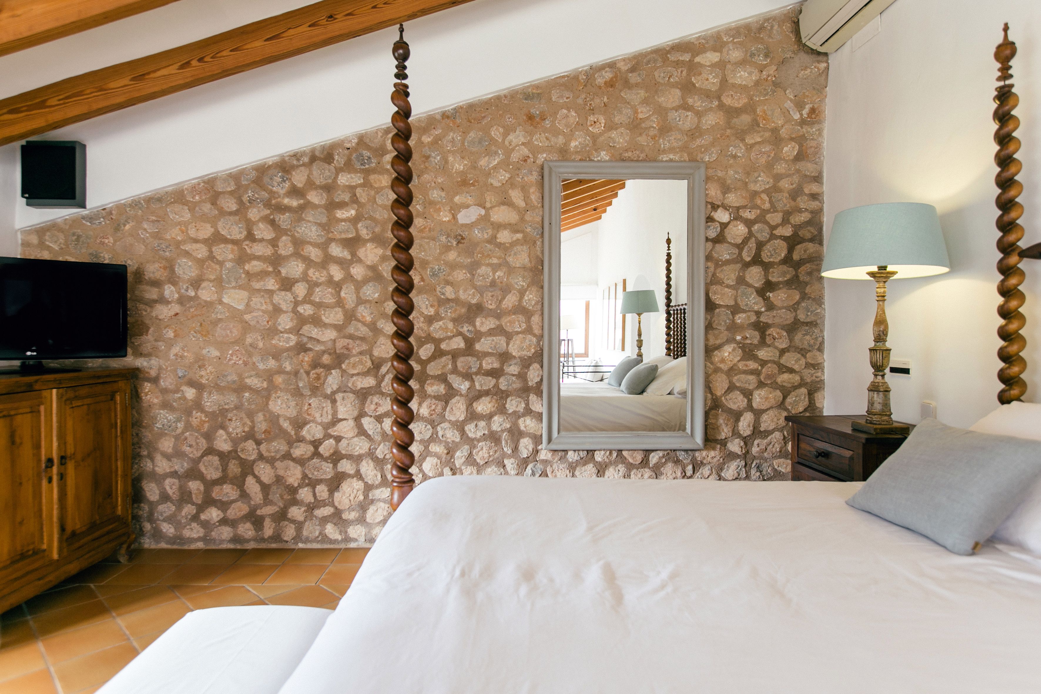 Un dormitorio con pared de piedra, cama con ropa de cama blanca, cabecero de madera, televisor sobre un mueble de madera, mesita de noche con lámpara y espejo en la pared.