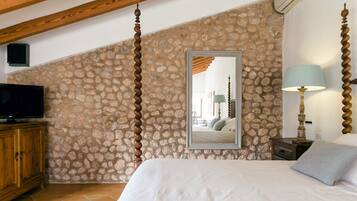 Un dormitorio con pared de piedra, cama con ropa de cama blanca, cabecero de madera, televisor sobre un mueble de madera, mesita de noche con lámpara y espejo en la pared.