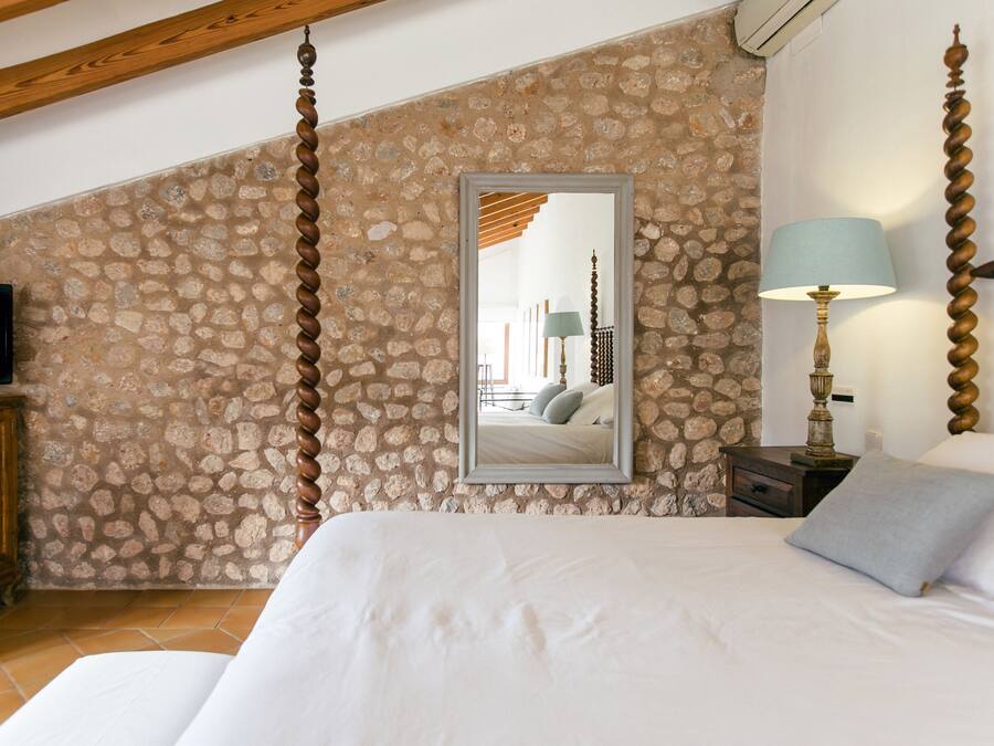 Un dormitorio con pared de piedra, cama con ropa de cama blanca, cabecero de madera, televisor sobre un mueble de madera, mesita de noche con lámpara y espejo en la pared.