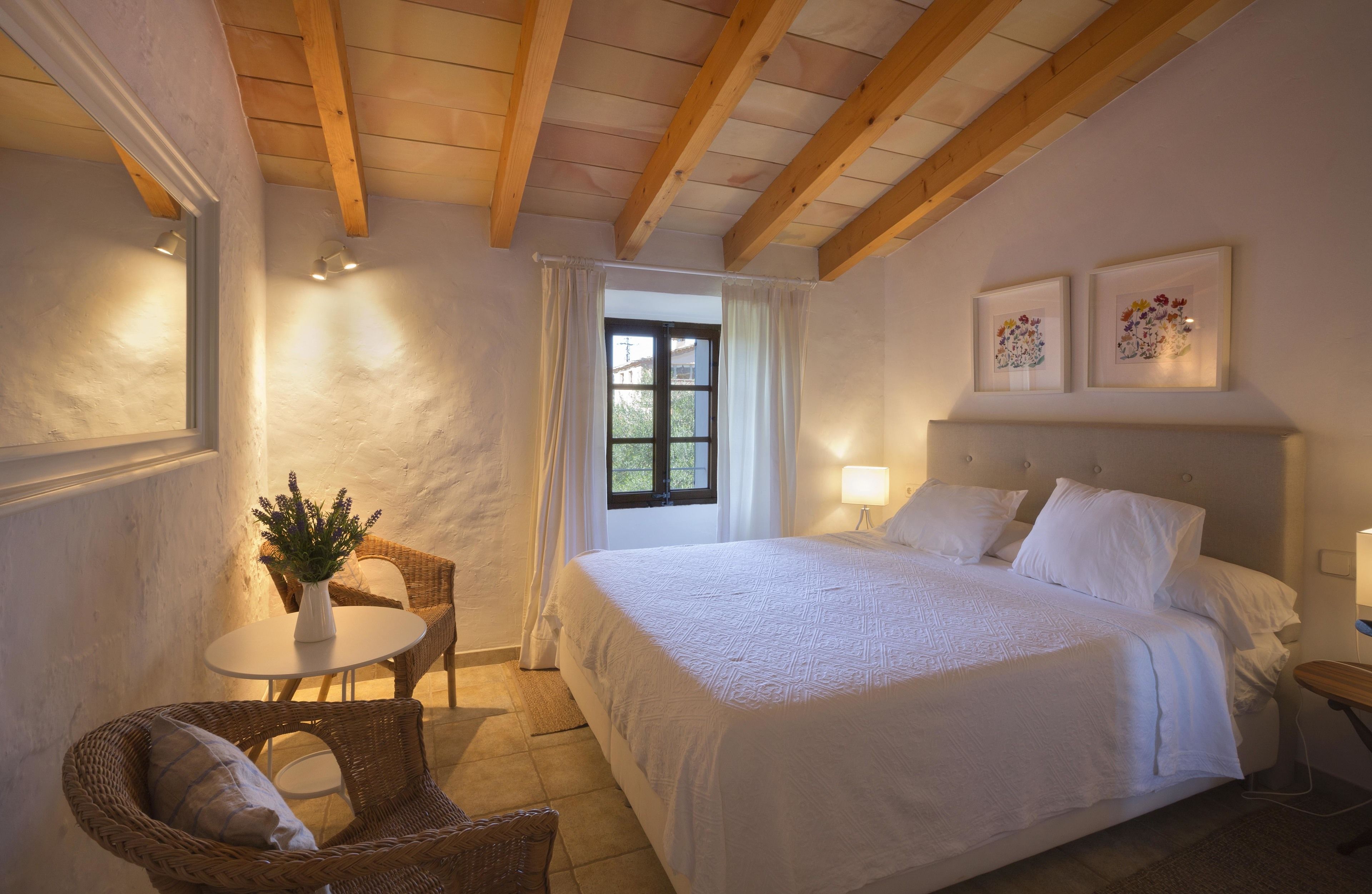 A bedroom with a large bed, a small table with a vase, wicker chairs, and a window with curtains.