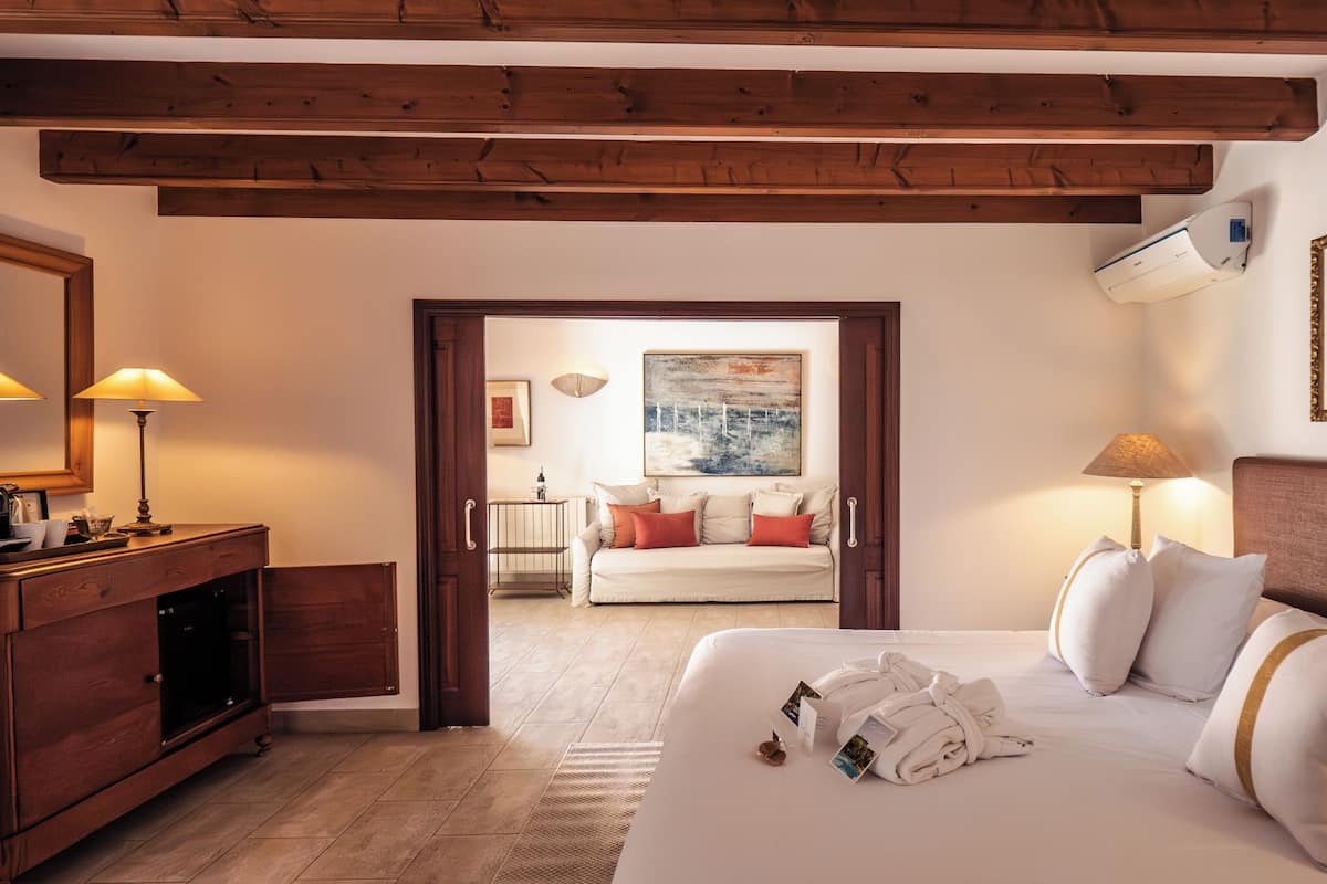 A hotel room with a bed, bedside lamps, a wooden ceiling, a painting, and a view through an open door.