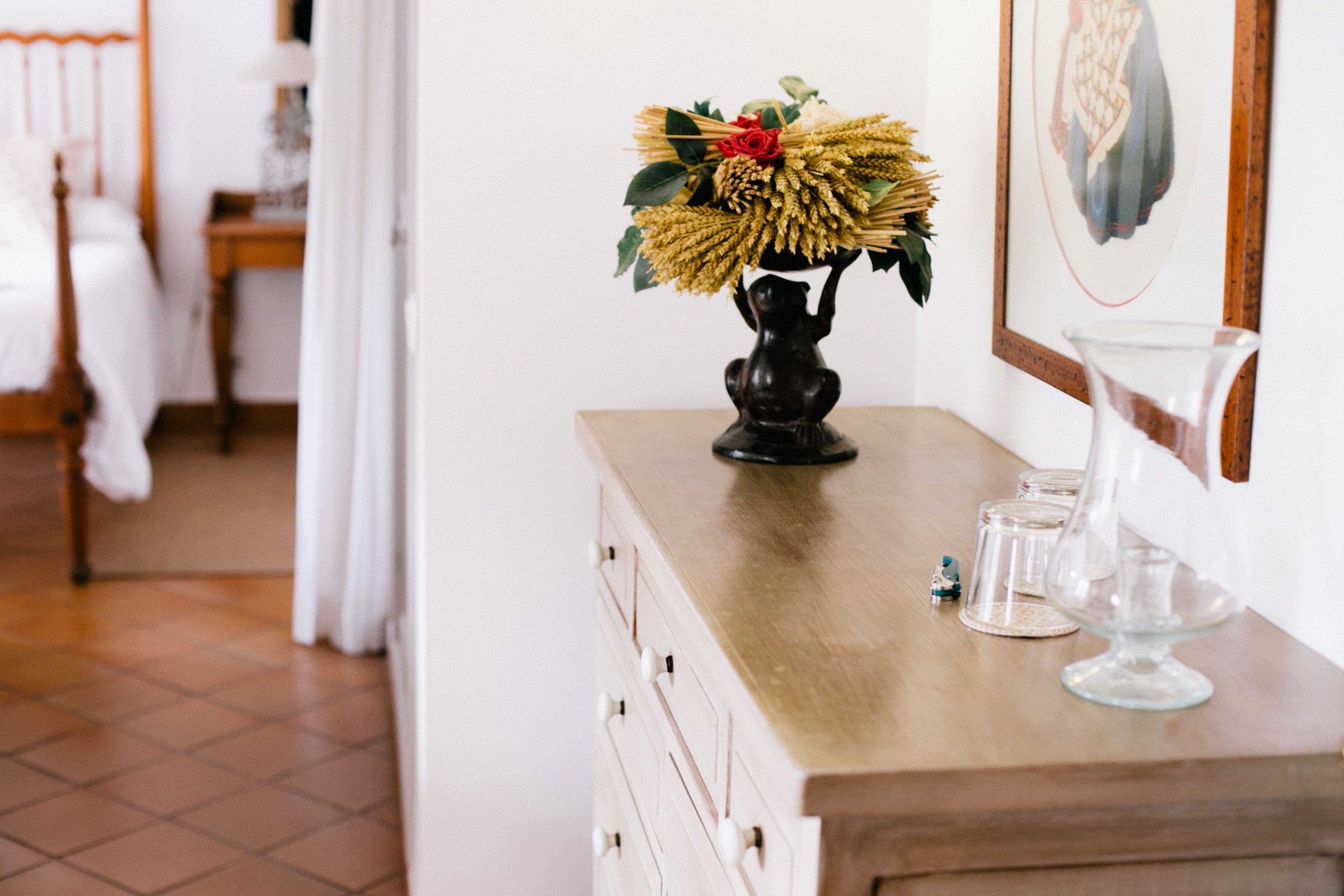 Et trækommode med en vase blomster, glas og et billede på væggen.