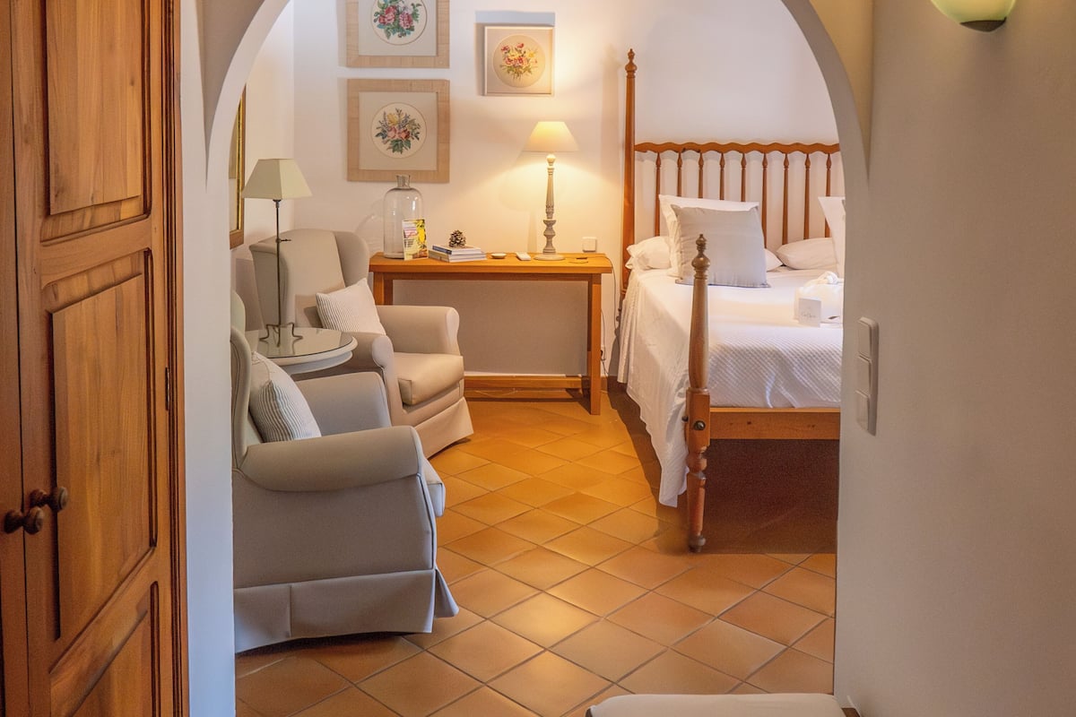 A bedroom with a wooden bed, a bedside table with a lamp, a chair, and a desk with a lamp. There are framed pictures on the wall and a tiled floor.