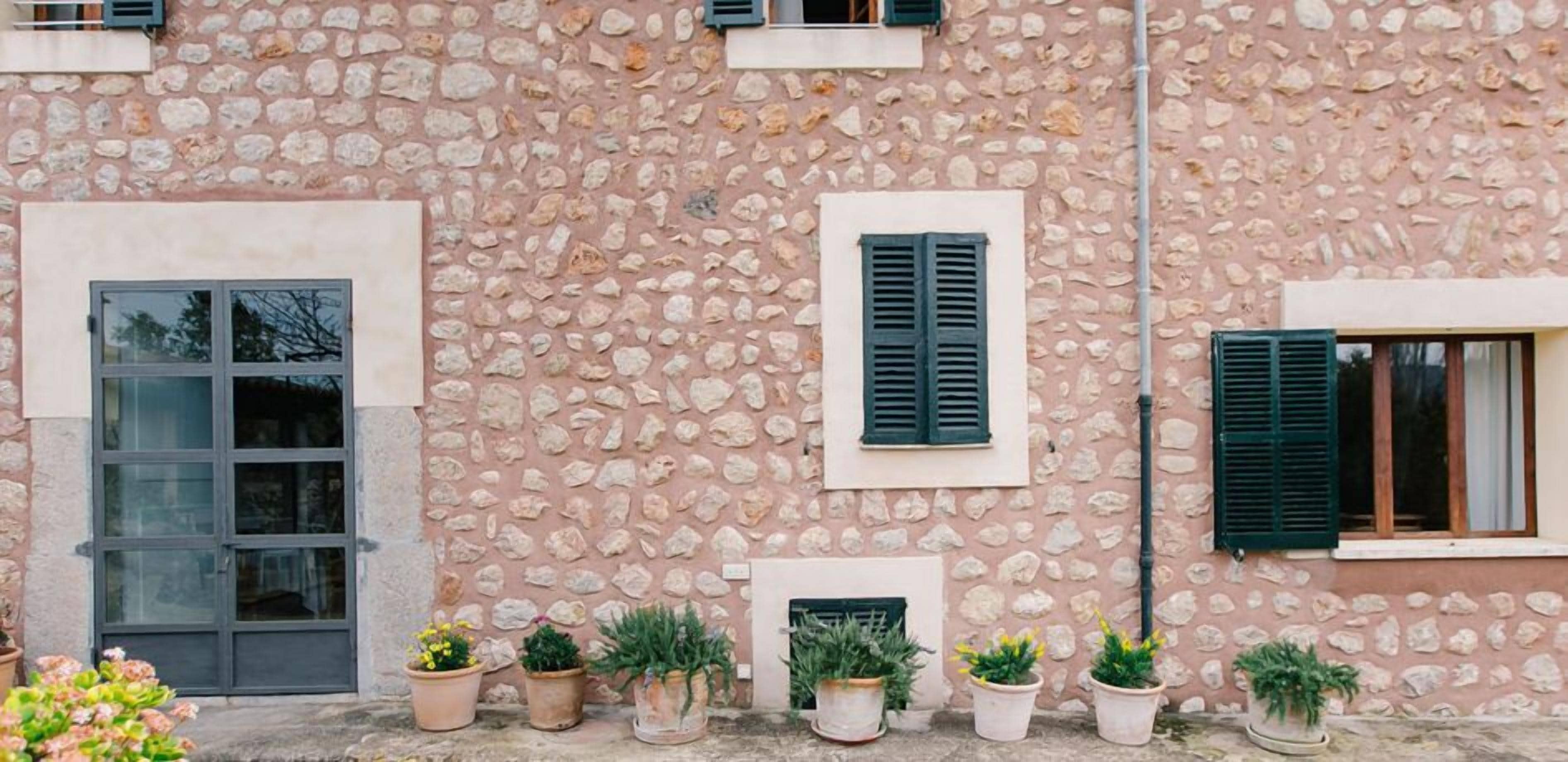 Un edificio de piedra con varias ventanas y puertas, plantas en macetas al frente y un muro de piedra.