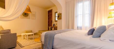 Una camera da letto con un letto grande, un divano, un tavolino e una sedia. Si vede una finestra con tende e una lampada a sospensione.