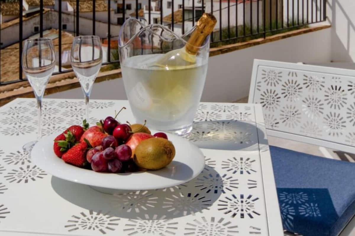 Una mesa con mantel blanco, un plato de fruta, dos copas de vino y una botella de champán en un balcón con vistas a un pueblo costero.