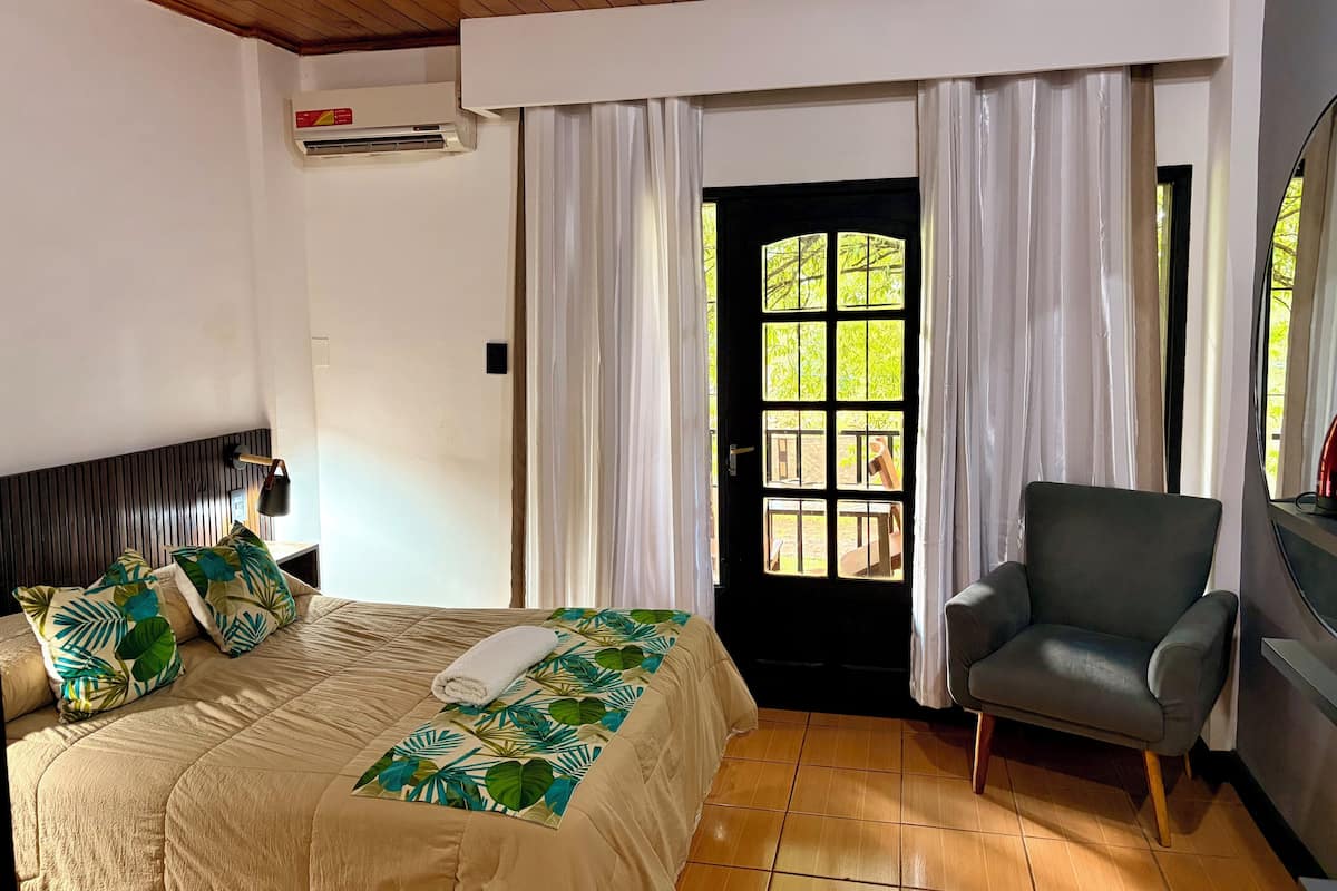 A hotel room with a bed, a chair, a window with curtains, and an air conditioning unit.