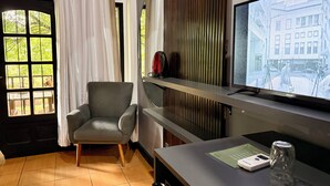 A hotel room with a large window, a grey armchair, a flat-screen TV on a stand, and a small table with a remote control and a glass.