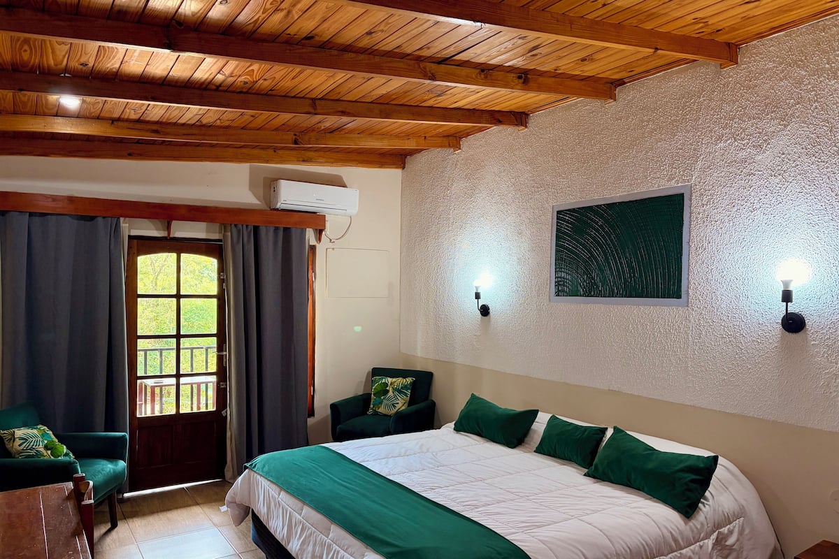 A bedroom with a wooden ceiling, a bed with green and white bedding, two wall-mounted lights, and a window with curtains.