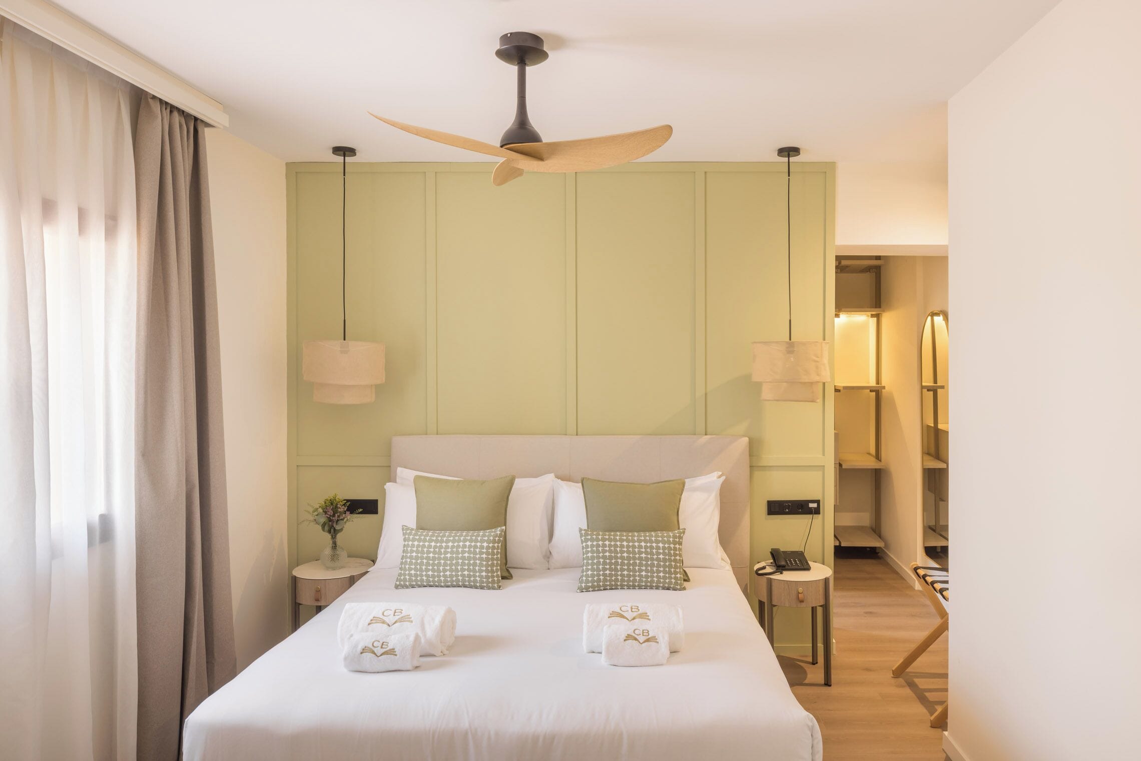 Quarto de hotel com cama, ventoinha no teto, luminárias de cabeceira, mesa de cabeceira e vista para o corredor.