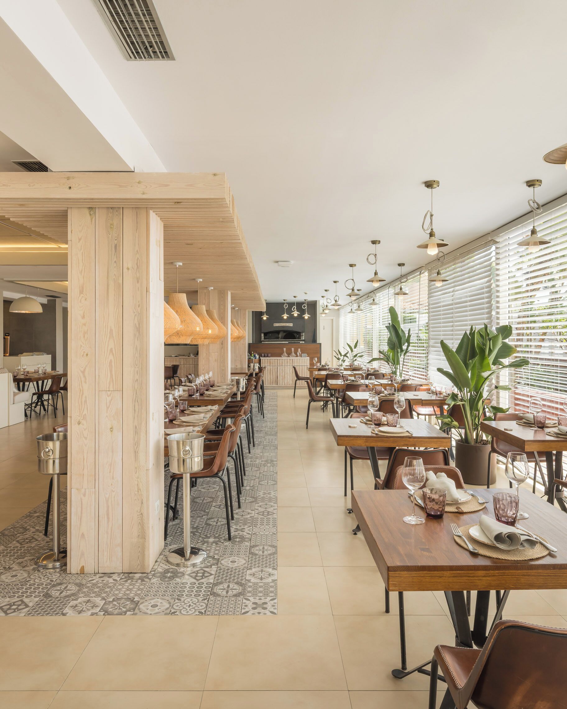 Un restaurant spacieux avec de hauts plafonds, des poutres en bois et de grandes fenêtres. Les tables sont dressées avec de la vaisselle et des verres, et on y trouve des plantes en pot.