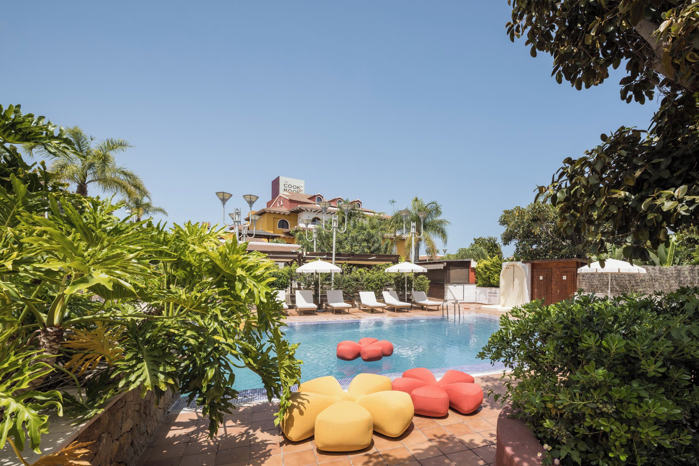 A pool area with lounge chairs, a sign reading 'Cook House,' and lush greenery.