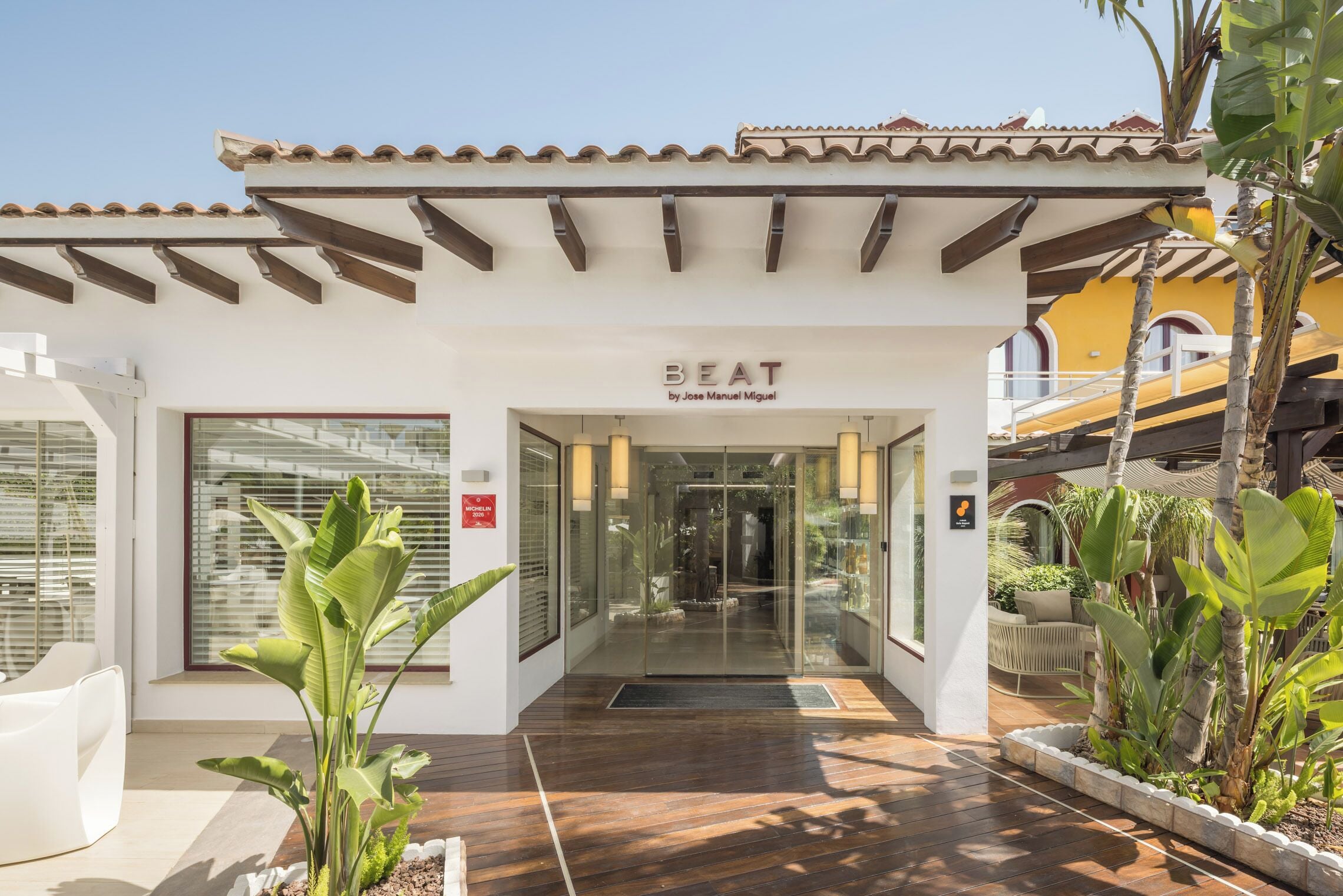 A building with a sign reading 'BEAT by Jose Manuel Miguel,' a glass entrance, and a wooden deck with plants.