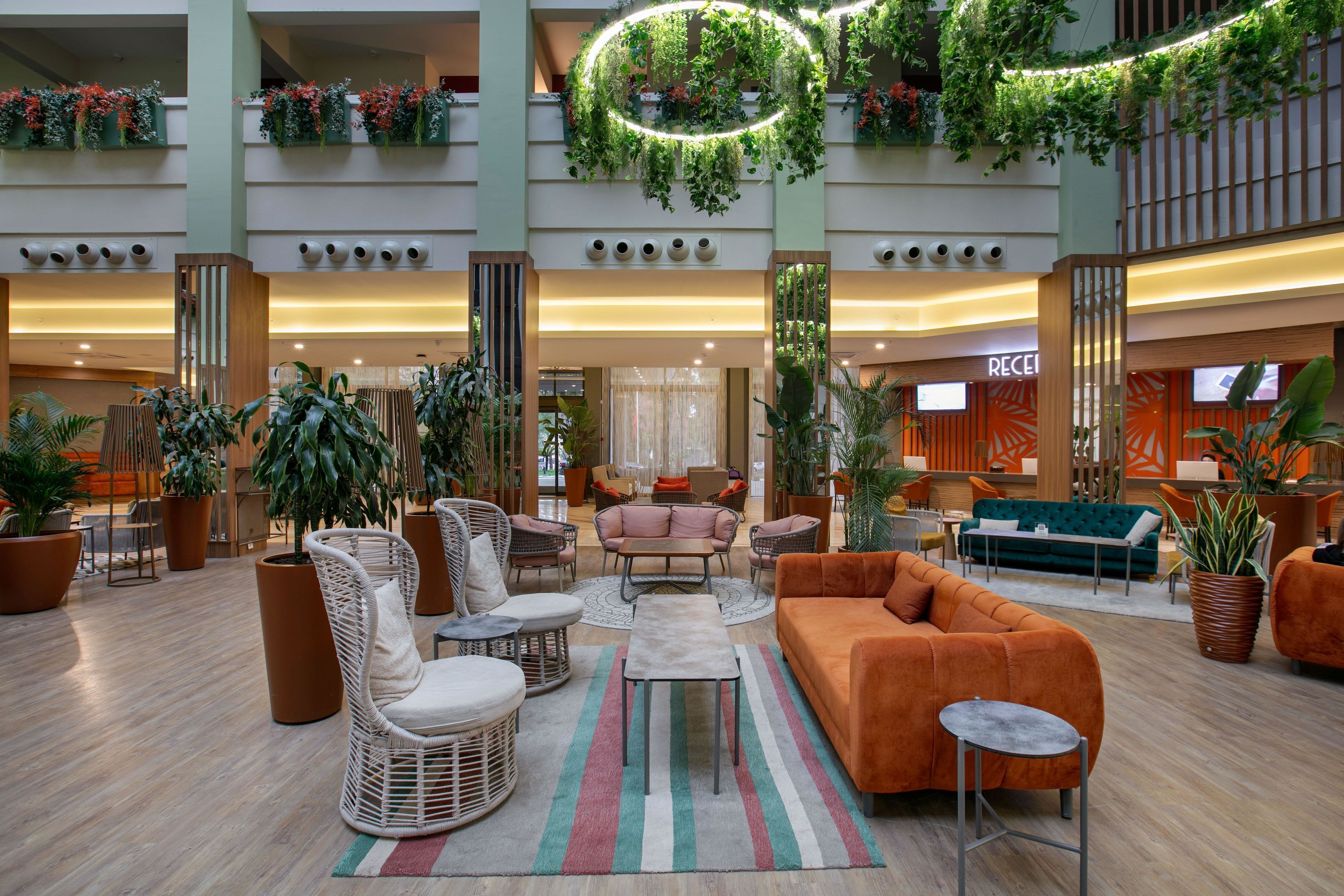 Un hall amplio con varias zonas de estar, incluyendo sofás y sillones, rodeado de grandes macetas con plantas y un mostrador de recepción central.