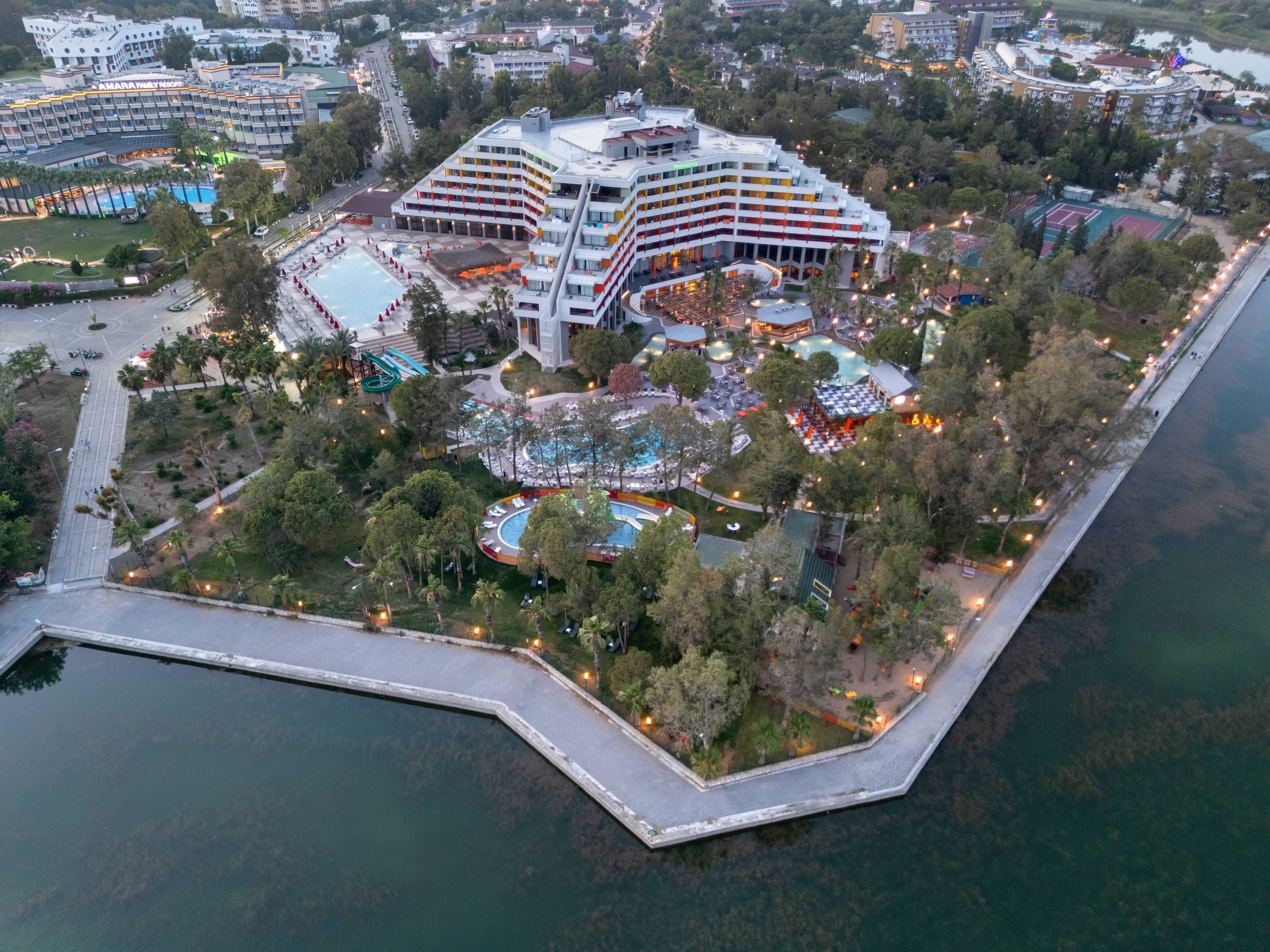 Et stort hotel med flere etager, en swimmingpool og et anlagt område med træer og en bro.