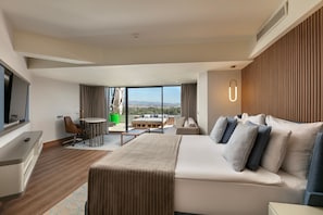 A modern hotel room with a large bed, a desk, a chair, a small table, and a balcony with a view. - Megasaray Resort Side (Manavgat)