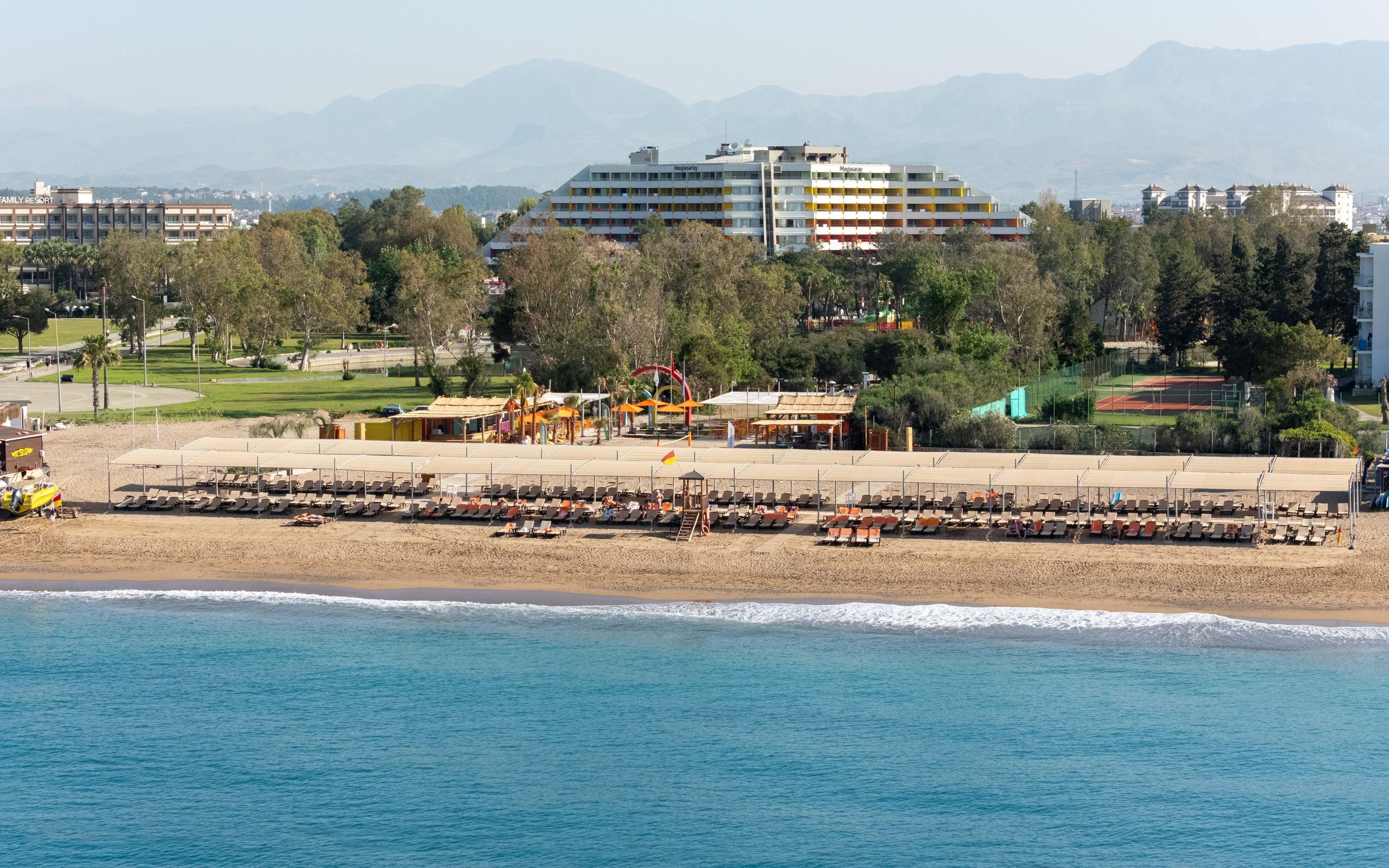 Un resort frente al mar con un edificio grande identificado como 'Megasary' al fondo, una playa de arena con tumbonas y vistas al océano.