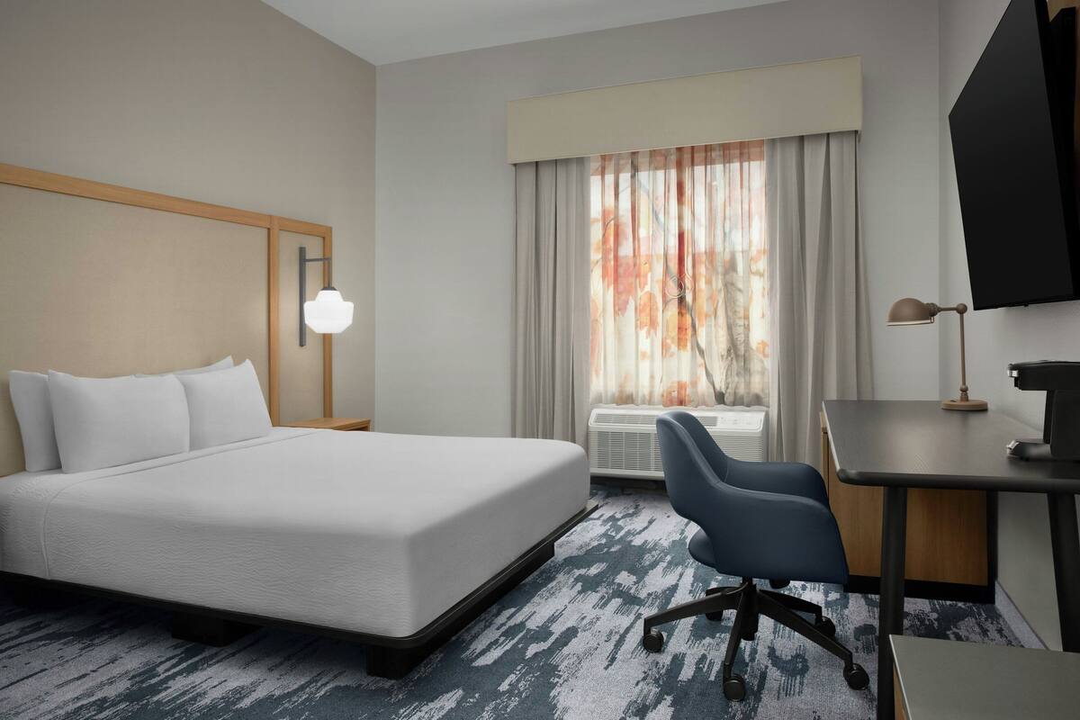 A hotel room with a bed, desk, chair, and a large window with curtains.