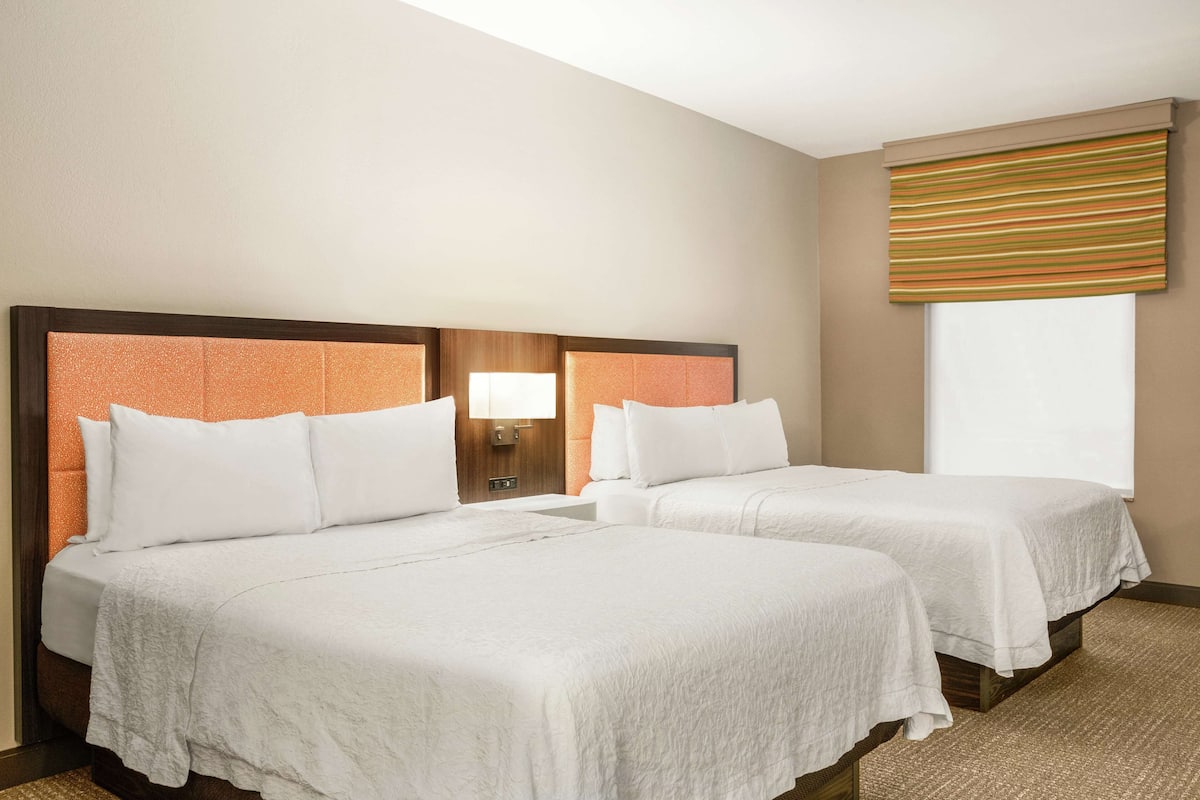 A hotel room with two beds, a nightstand with a lamp, a window with a striped blind, and a carpeted floor.