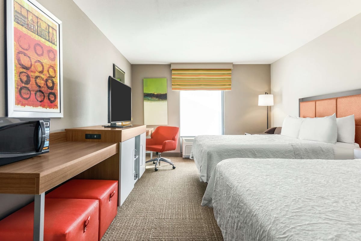 A hotel room with two beds, a desk with a TV, a red chair, and a window with blinds.