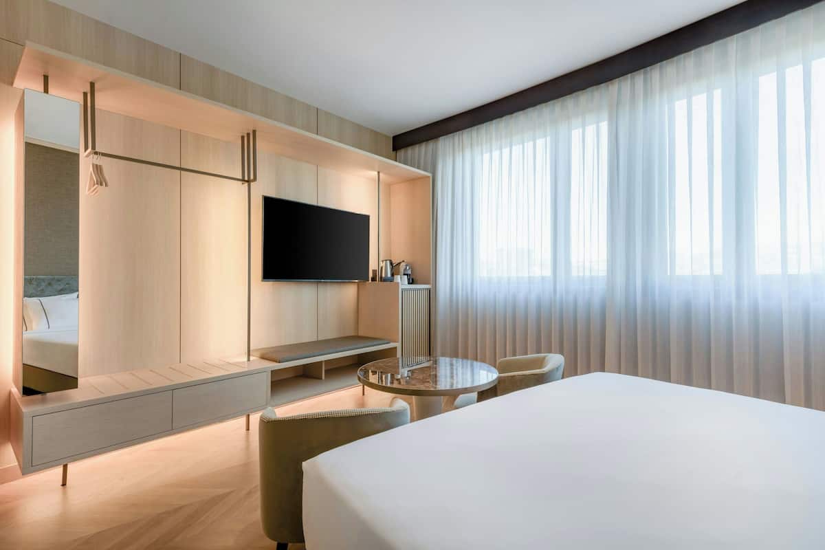 A modern hotel room with a large bed, a round dining table, a sofa, a TV mounted on a wall, and a large window with sheer curtains.
