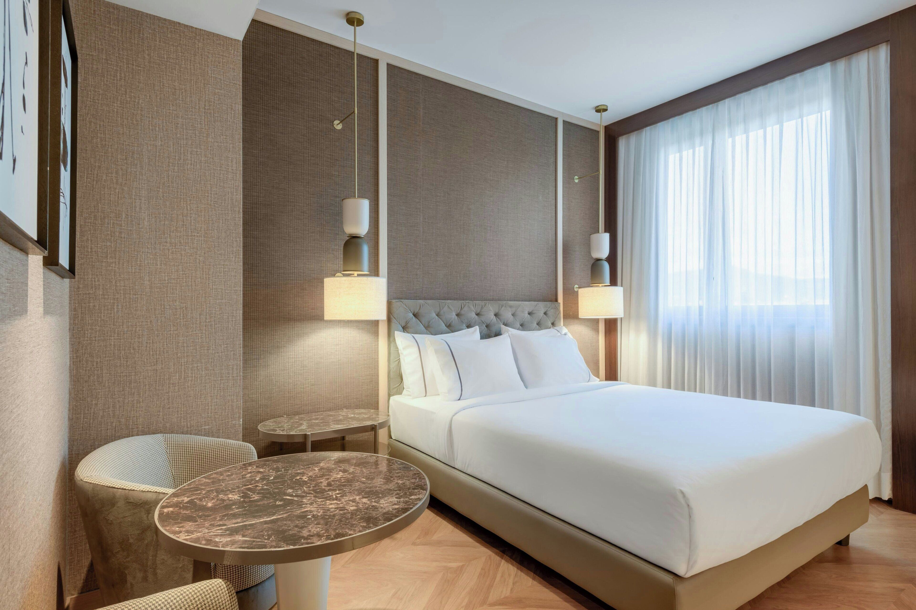 A hotel room with a large bed, two bedside tables, a round table, and a chair.