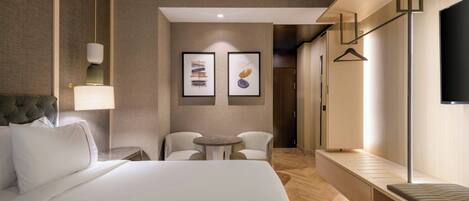 A modern hotel room with a large bed, a bedside table, a lamp, a small round table with two chairs, and a TV mounted on the wall.