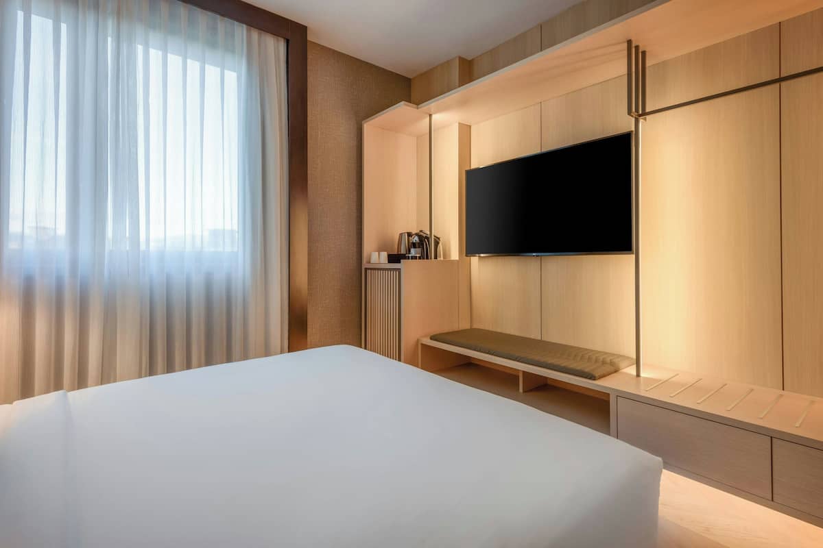 A hotel room with a large bed, a bench, a flat-screen TV mounted on a wooden wall, and a window with sheer curtains.