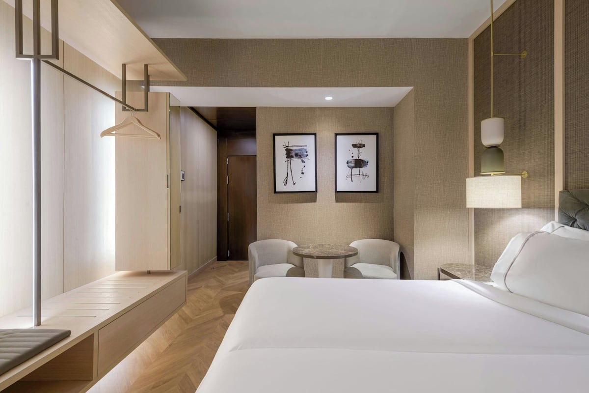A hotel room with a large bed, a bedside table, a small round table with two chairs, a hanging coat rack, and two framed artworks on the wall.