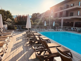 Zona junto a la piscina con tumbonas y sombrillas, una terraza de madera y un edificio moderno al fondo.