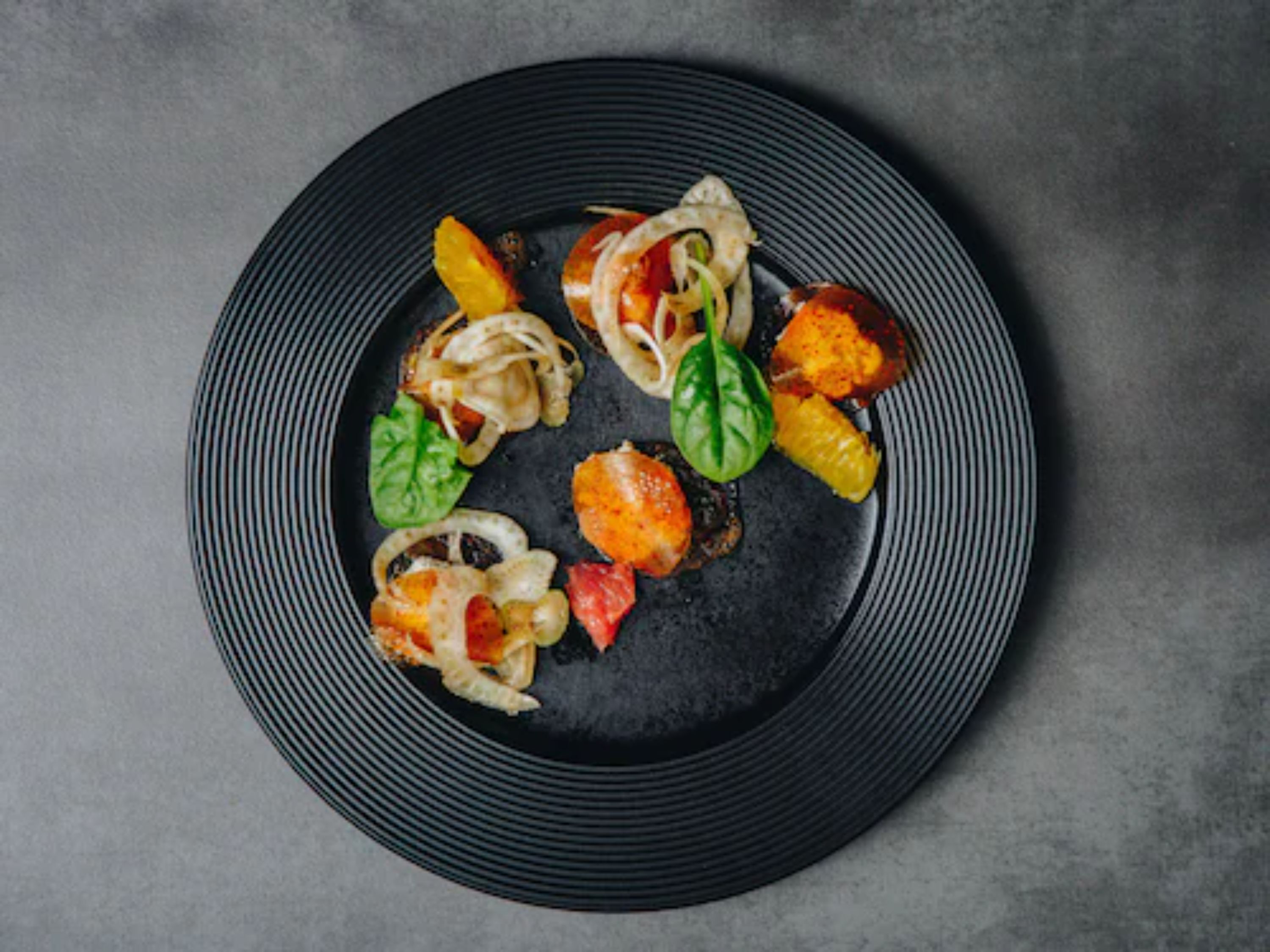 Un plato negro con diversos alimentos, entre ellos camarones, verduras y posiblemente pasta.