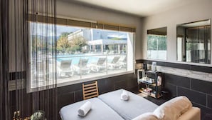 Quarto de hotel com cama, uma mesinha, prateleira com artigos de higiene e vista para a piscina e um edifício lá fora.