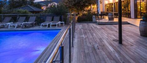 Una terraza de madera con piscina, sillas de descanso y un edificio con área de estar al aire libre.