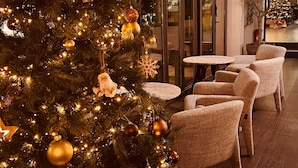 Árvore de Natal com enfeites e luzes no hall de um hotel, com área de estar e porta de vidro.