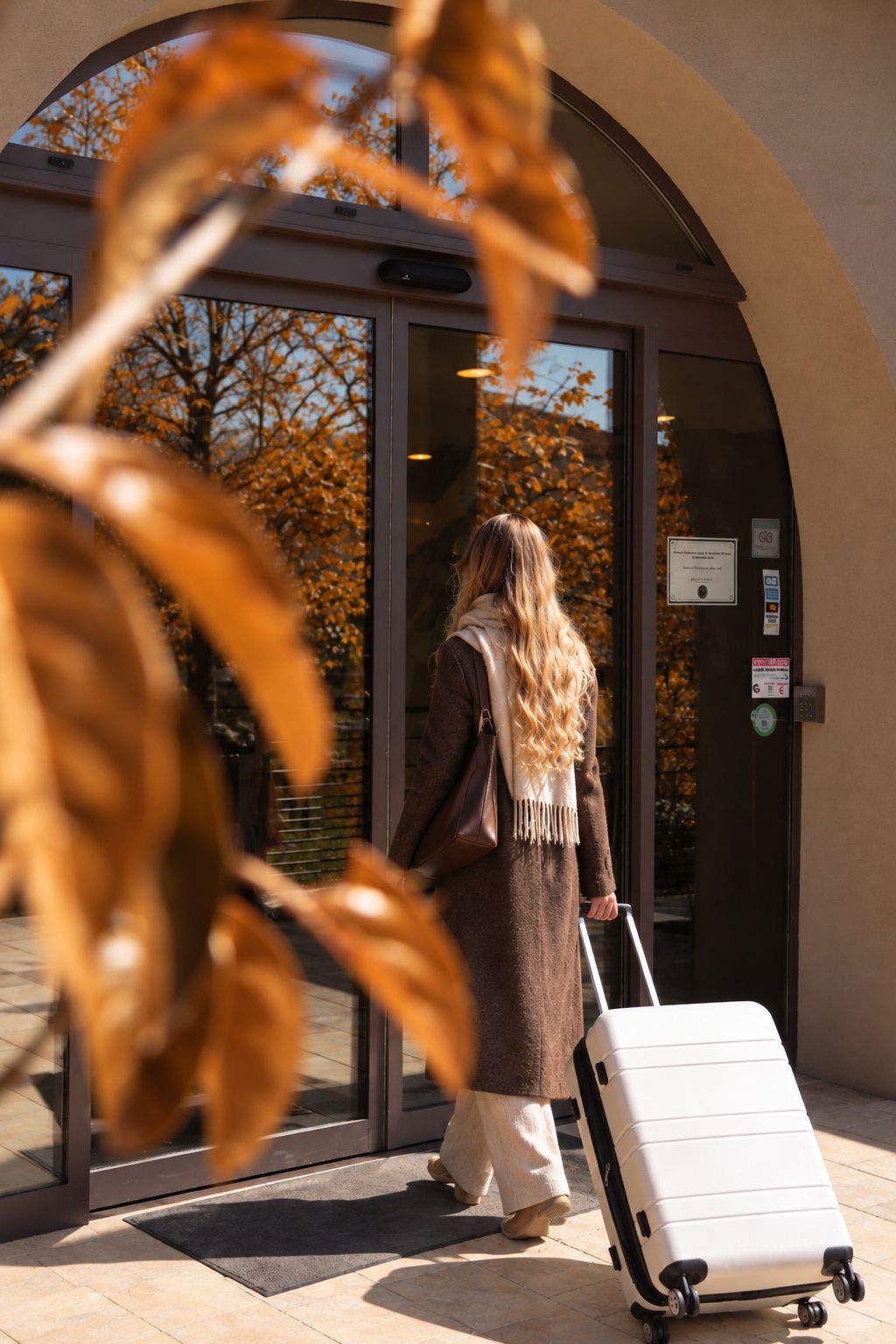 A person exiting a building with a suitcase, through a glass door with a view of trees.