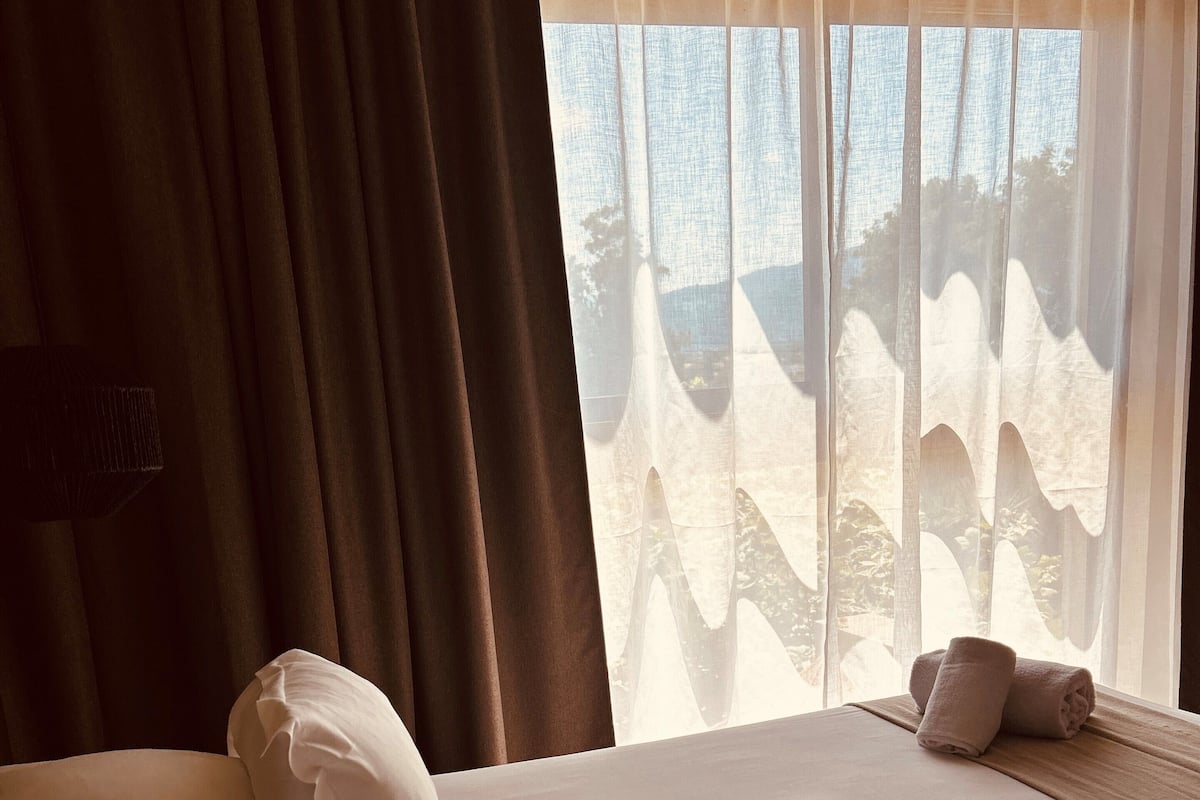 Une chambre d’hôtel avec un lit, des oreillers, une fenêtre avec des rideaux légers et une vue sur les arbres à l’extérieur.