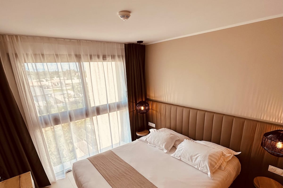 Une chambre d’hôtel avec un grand lit, une table de chevet, une commode et une vue sur l’extérieur à travers des rideaux transparents.